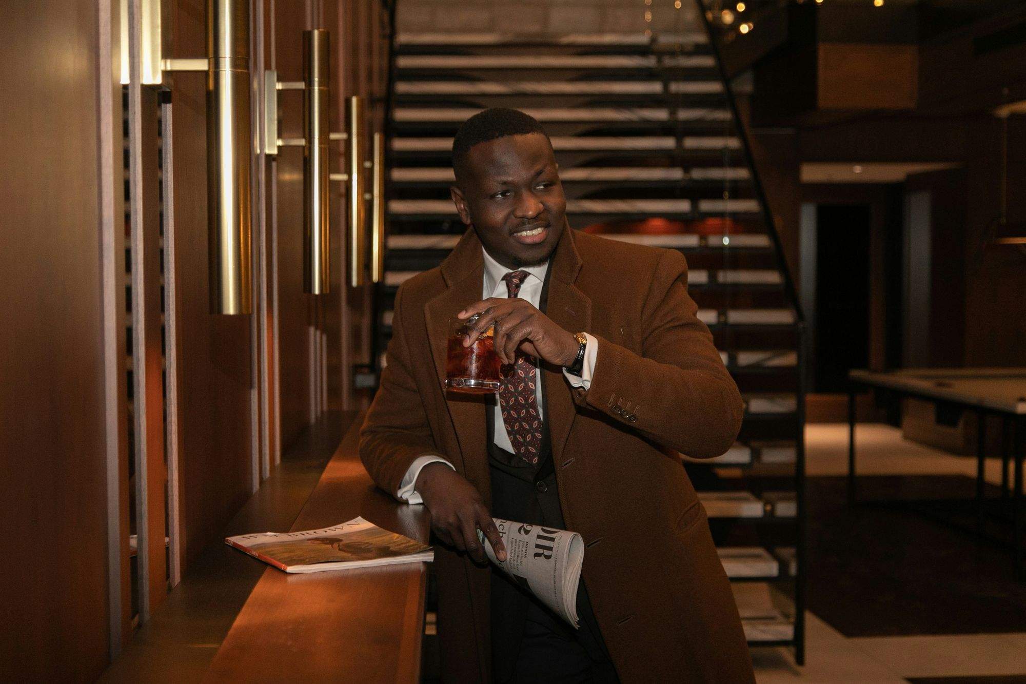 A smiling man in a camel coat holding a drink and a newspaper